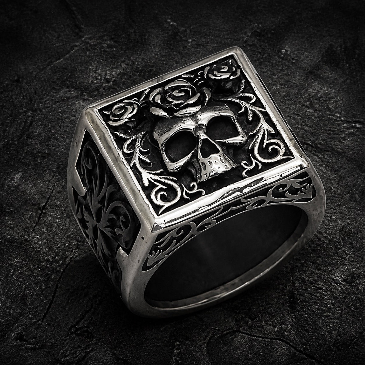 A silver-toned gothic ring featuring a square face with an embossed skull, roses, and ornate scrollwork, photographed on a dark textured surface.