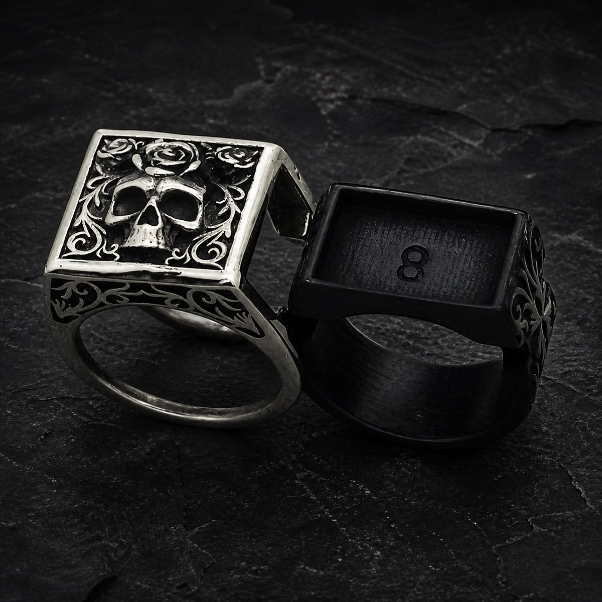 Two gothic-style rings displayed on a dark textured slate background. The ring on the left is made of oxidized silver with a square face featuring a detailed skull and rose motif.