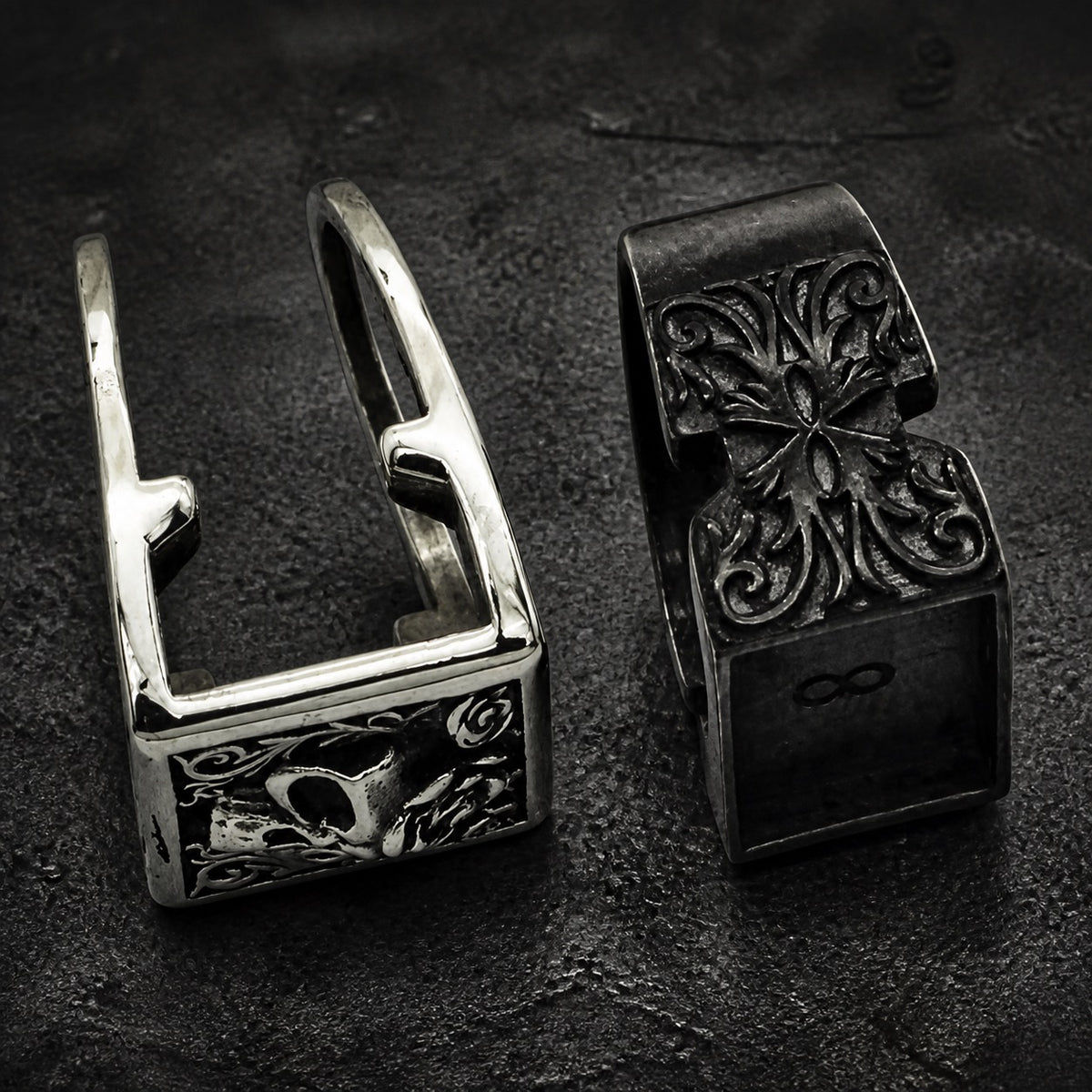 Two gothic-style metal rings displayed side-by-side on a dark textured surface. The left ring is silver-toned with a skull and floral engraving.