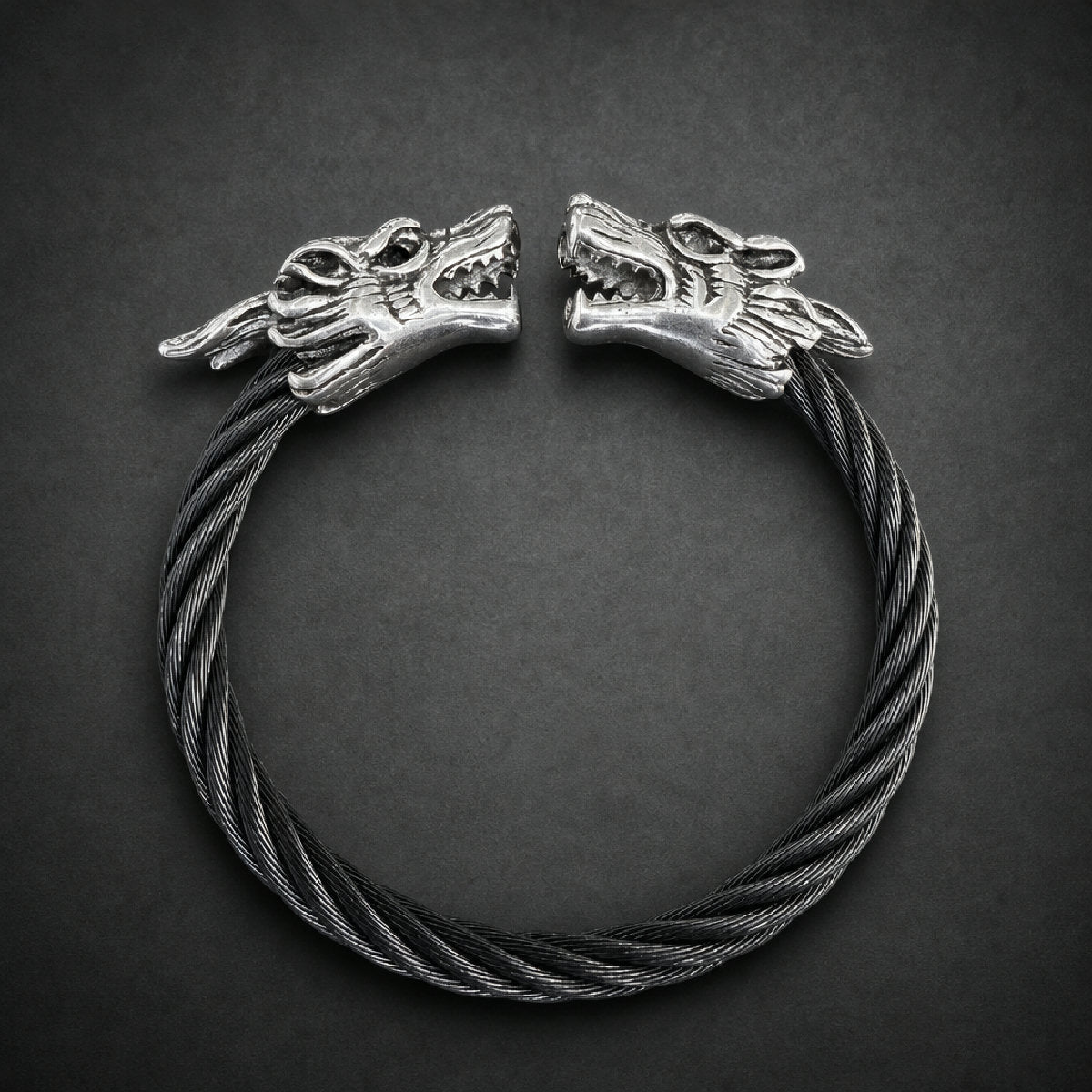 Dual snarling wolf head bracelet with a twisted dark cable band made of 316L surgical stainless steel on a wooden surface.
