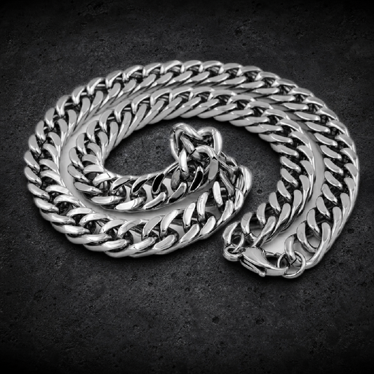 Highly polished 316L stainless steel curb chain necklace, featuring thick, flattened interlocking links. The chain is coiled on a white surface, showcasing its robust construction and shiny finish, with a lobster clasp visible at one end.