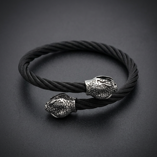 Coiled black stainless steel cable bracelet with two silver or white steel serpent heads facing each other, detailed scales, minimalist background.